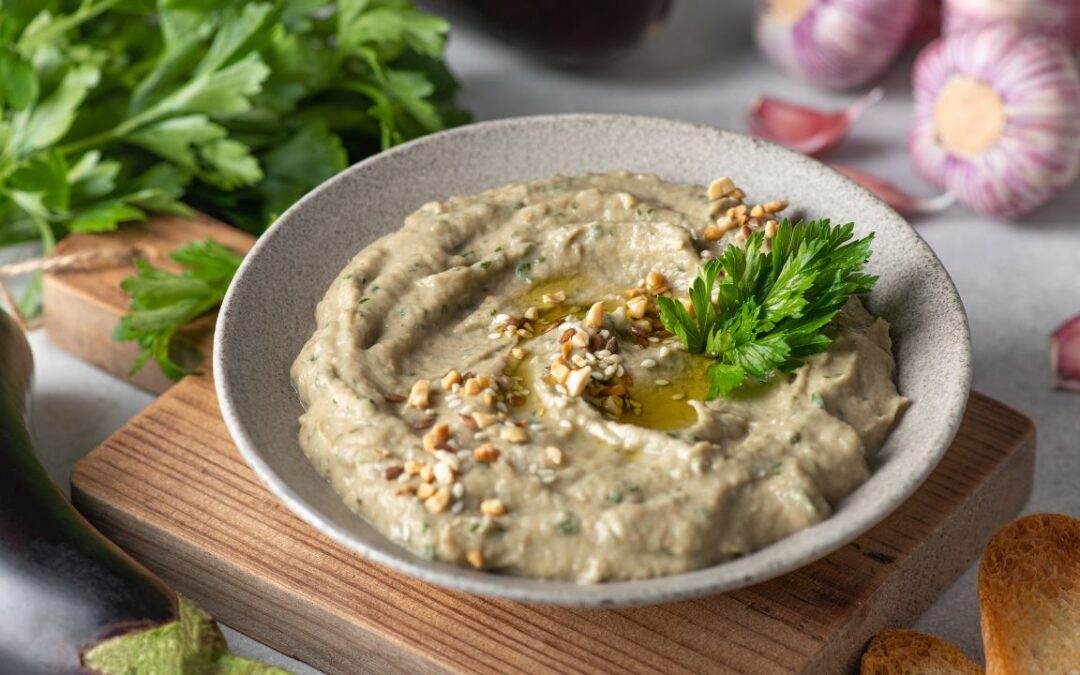 A bowl of baba ganoush topped with chopped nuts, olive oil, and a sprig of parsley, served on a wooden board with slices of toasted bread on the side. Fresh vegetables, including eggplant and garlic, are visible in the background