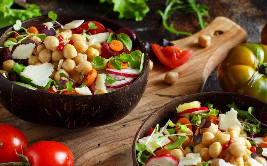 A vibrant chickpea salad served in a dark wooden bowl. The salad is a colorful mix of plump chickpeas, red radish slices, orange carrot rounds, deep red beet chunks, fresh green lettuce, and sprinkles of pomegranate seeds.
