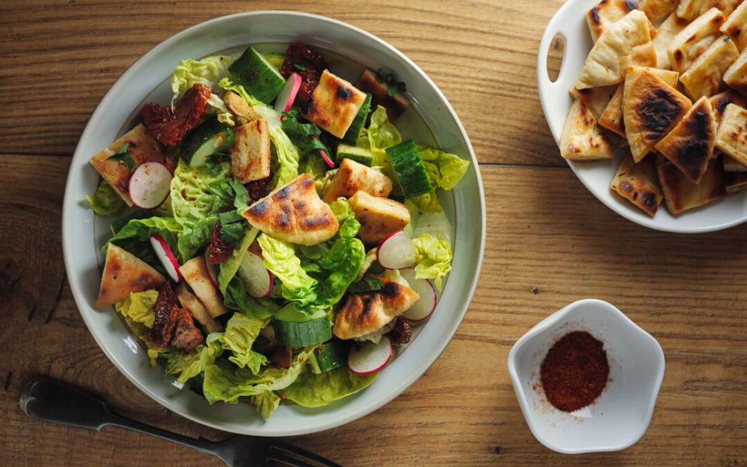A bowl of Fattoush salad on a wooden table, featuring mixed greens, radishes, grilled pita bread, and sun-dried tomatoes