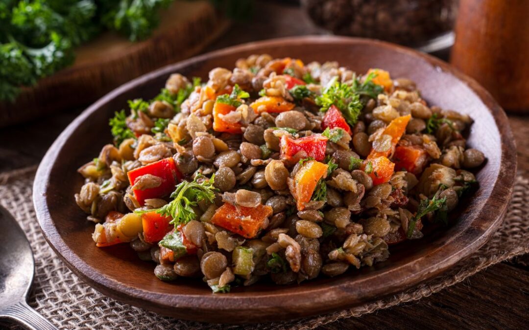A close-up of a Lebanese-style lentil salad served in a rustic wooden bowl. The salad is a mix of cooked brown lentils, diced red and orange bell peppers, chopped parsley, and other herbs.