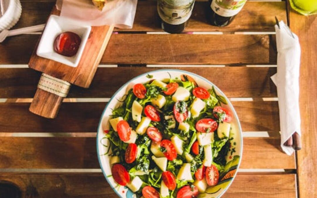 A fresh, vibrant bowl of mediterranean diet salad on a wooden table, featuring a mix of greens, cherry tomatoes cut in halves, and chunks of white cheese, possibly mozzarella.