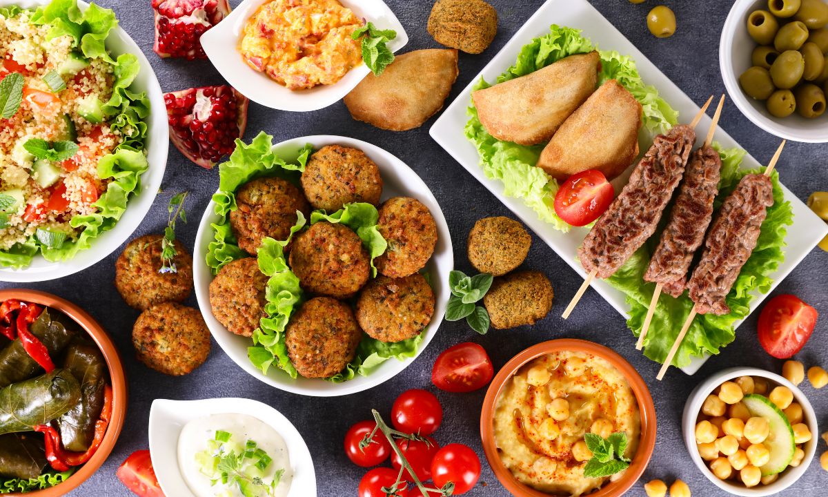 An assortment of Mediterranean diet dishes displayed on a table, including falafel, stuffed grape leaves, grilled meat skewers, various dips like hummus and tzatziki, fresh salad with quinoa, and ripe tomatoes, with a side of olives.