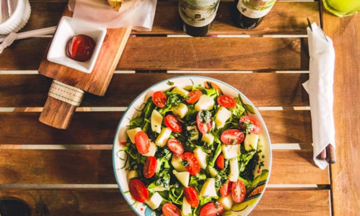 A fresh, vibrant bowl of mediterranean diet salad on a wooden table, featuring a mix of greens, cherry tomatoes cut in halves, and chunks of white cheese, possibly mozzarella.