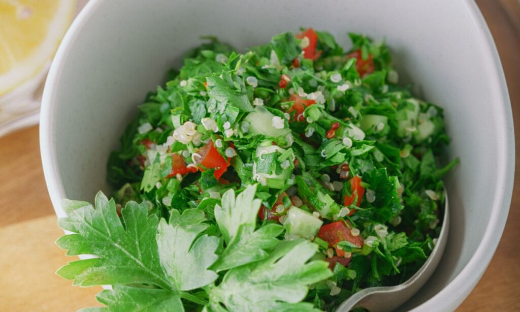 Close-up of tabouli in a white bowl with a fresh parsley leaf garnish, highlighting the green herbs, diced tomatoes, and scattered quinoa grains