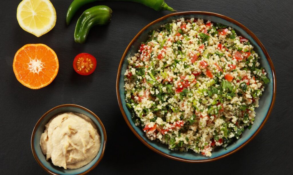 A bowl of tabouli salad, garnished with fresh parsley, diced tomatoes, and green onions with a small bowl of hummus.