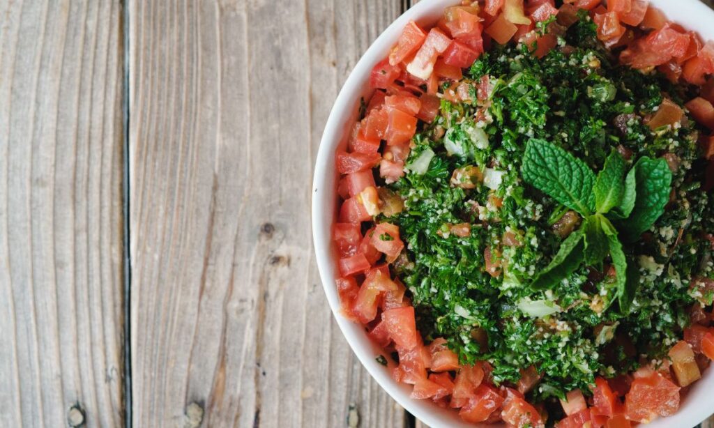 Close-up of a bowl of tabouli salad topped with a sprig of mint, with finely chopped parsley and diced tomatoes, set on a wooden table