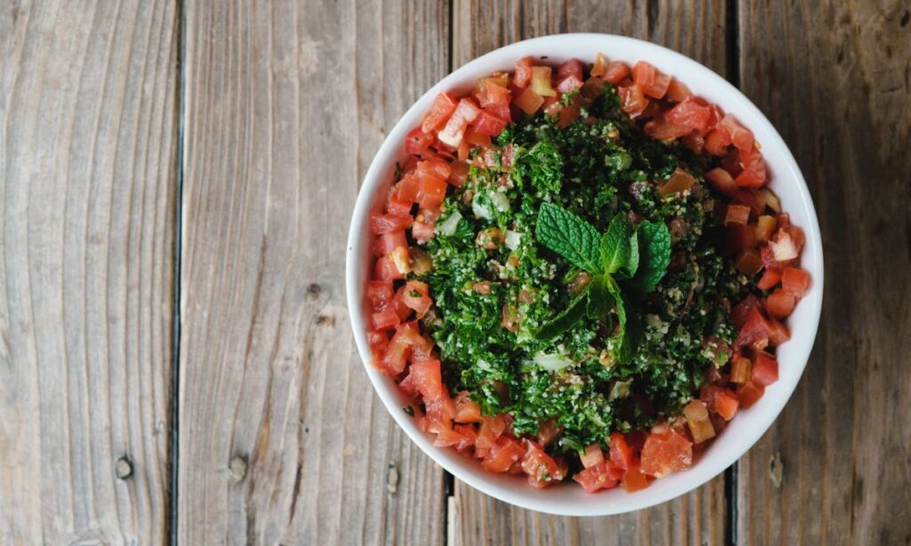 A bowl of tabouli salad centered on a rustic wooden table, topped with a sprig of mint, highlighting the dish's fresh herbs and diced tomatoes