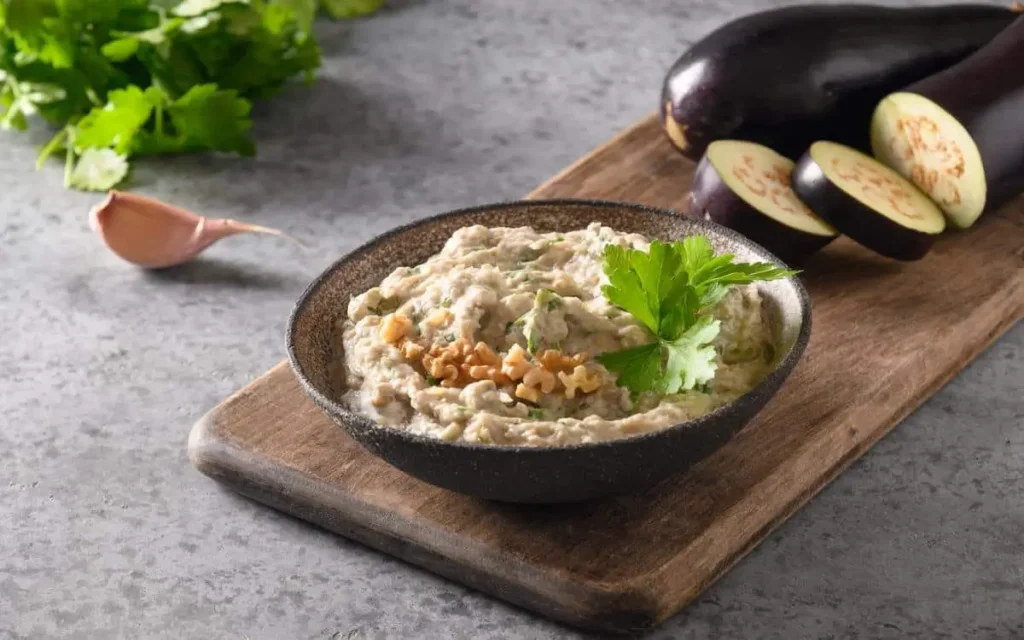 A bowl of creamy baba ganoush garnished with fresh parsley and walnuts, served on a wooden board with eggplants and garlic