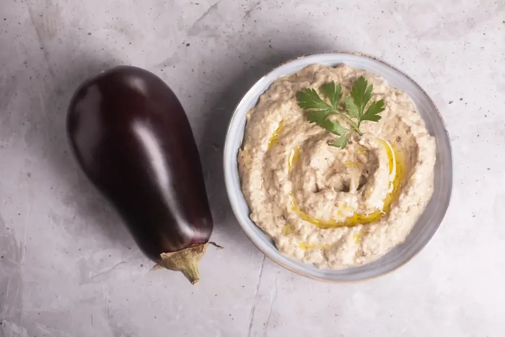 Freshly prepared baba ganoush in a bowl garnished with olive oil and parsley, next to a whole eggplant on a marble background.