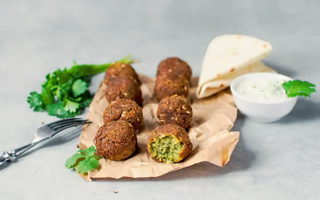 Freshly made falafel balls on parchment paper, served with a bowl of yogurt sauce, flatbread, and parsley garnish on a gray background