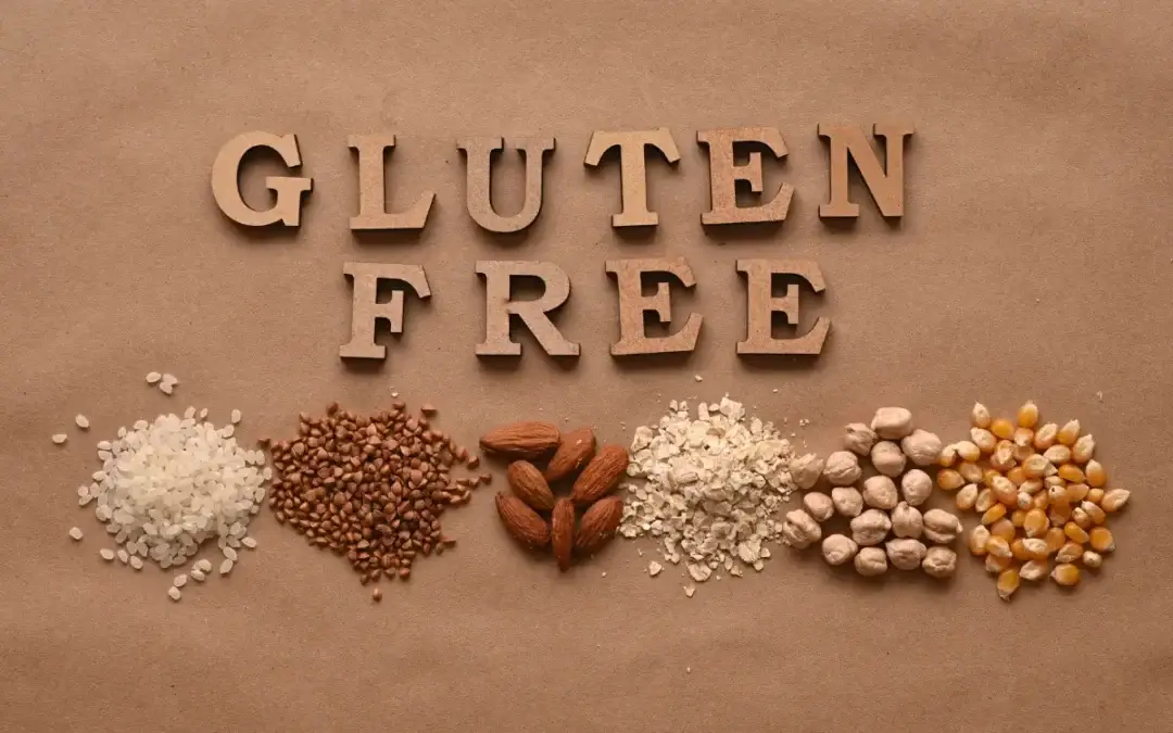 Gluten-free spelled out with wooden letters, surrounded by gluten-free foods such as rice, buckwheat, almonds, oats, chickpeas, and corn on a brown background