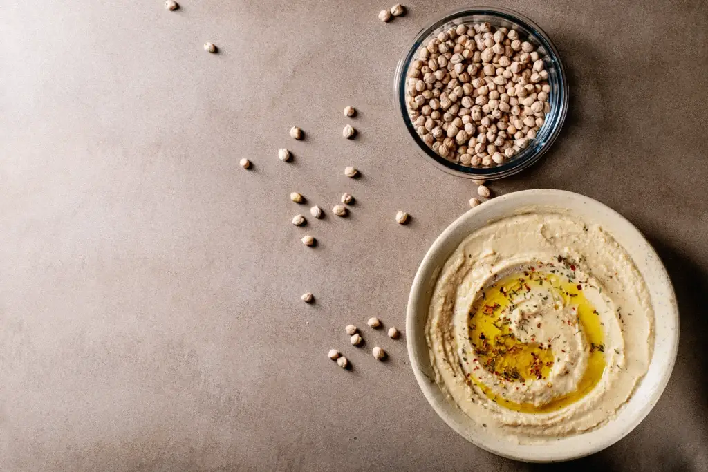 Traditional hummus served in a bowl drizzled with olive oil and spices, surrounded by whole chickpeas on a beige background.