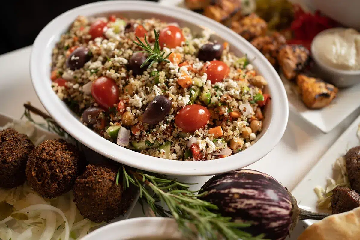 A quinio Salad in a white oval bowl, the quinio salad is mixed with sliced olives, cherry tomatoes, and a sprinkle of chopped parsley. it is situated in an 30 degreen inclination to the NW of the picture and there is a ros,erry leave with an exotic eggplant fresh vegetables