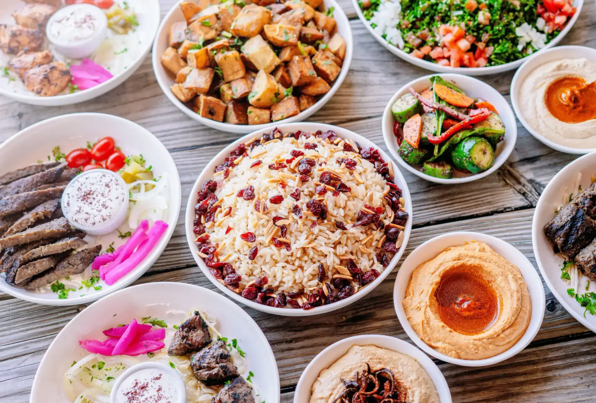 A spread of middle eastern food which is served by Aladdin Mediterranean Grill and the spread is sliced lamb Gyro, almond cranberry rice,lebanese cucumber salad, steak beef filet kebab, chipotle chicken kebab, and tabouli salad all served on a rustic wooden table.