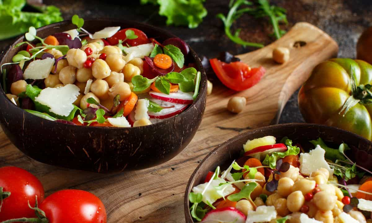 A vibrant chickpea salad served in a dark wooden bowl. The salad is a colorful mix of plump chickpeas, red radish slices, orange carrot rounds, deep red beet chunks, fresh green lettuce, and sprinkles of pomegranate seeds.