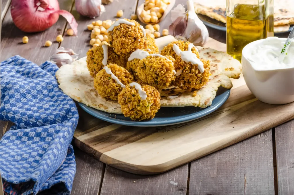 A beautifully arranged plate of crispy gluten-free falafel balls with one broken open to reveal a green herbed center, accompanied by parsley, flatbread, and a creamy yogurt sauce, set against a neutral gray surface