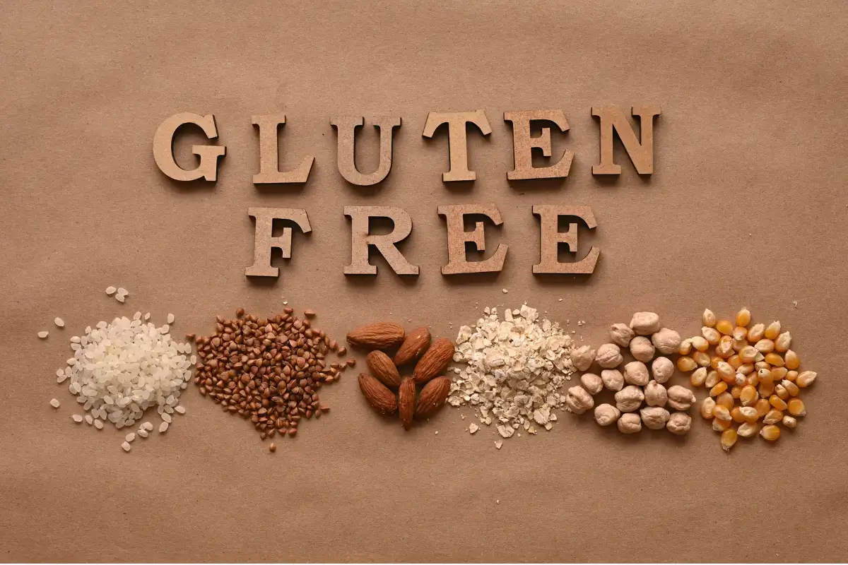 Gluten-free spelled out with wooden letters, surrounded by gluten-free foods such as rice, buckwheat, almonds, oats, chickpeas, and corn on a brown background