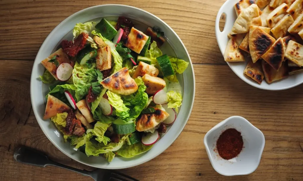 A bowl of Fattoush salad on a wooden table, featuring mixed greens, radishes, grilled pita bread, and sun-dried tomatoes