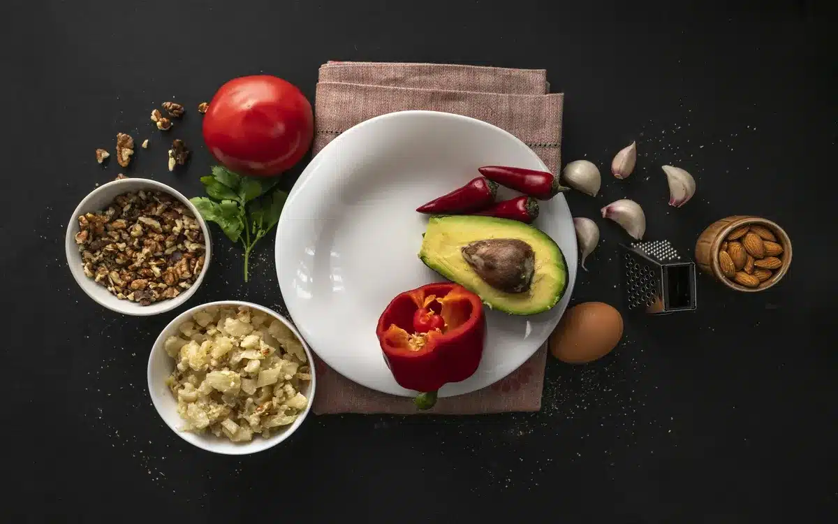 Keto Mediterranean ingredients including avocado, walnuts, red peppers, almonds, garlic, and herbs on a dark background.