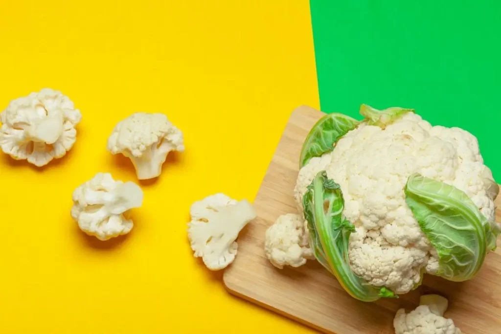 Whole cauliflower head and loose florets on a cutting board against bright yellow‑and‑green backdrop.